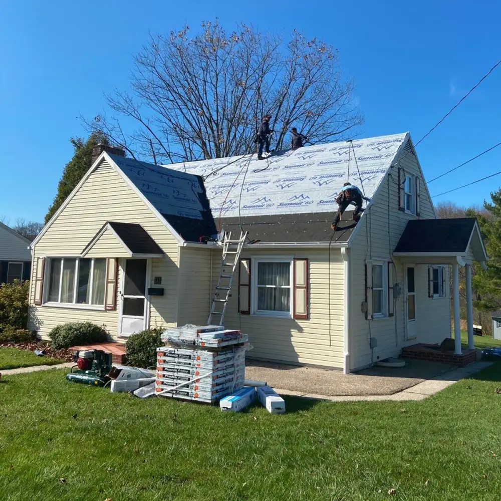 A crew on a yellow Cape Cod roof laying new underlayment, with shingle bundles on the lawn