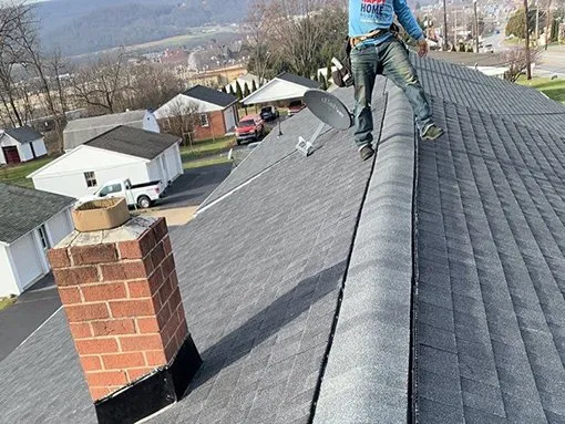 A roofer walking the ridge of a new grey roof beside a brick chimney, the valley town behind